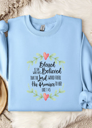 A light blue sweatshirt with a printed design and text, placed on a bed with a white blanket and a white mug with a heart design.