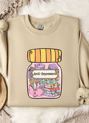 a beige sweatshirt with a colorful graphic design depicting a jar with various objects inside, including a book and a flower. The sweatshirt is placed on a bed with a white blanket and a white mug with a heart design.