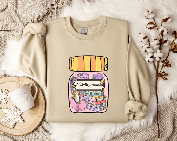 a beige sweatshirt with a colorful graphic design depicting a jar with various objects inside, including a book and a flower. The sweatshirt is placed on a bed with a white blanket and a white mug with a heart design.
