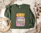 a green sweatshirt with a colorful graphic design depicting a jar with various objects inside, including a book and a flower. The text on the sweatshirt reads "Ani-Depressants".
