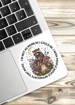 A sticker with a raccoon sitting on a mushroom and surrounded by flowers and plants is placed on a laptop keyboard.