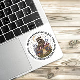 A sticker with a raccoon sitting on a mushroom and surrounded by flowers and plants is placed on a laptop keyboard.