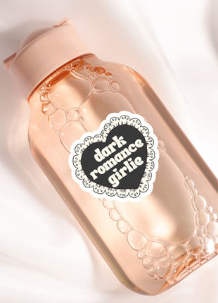 A pink bottle with a black heart-shaped label that says "Dark Romance Girlie" on it.