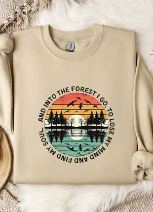 a beige sweatshirt with a graphic design depicting a sunset over a forest, along with the text "And into the forest go I lose my soul" printed on it. The sweatshirt is laid out on a bed with a white blanket, and there are some flowers and a mug nearby.