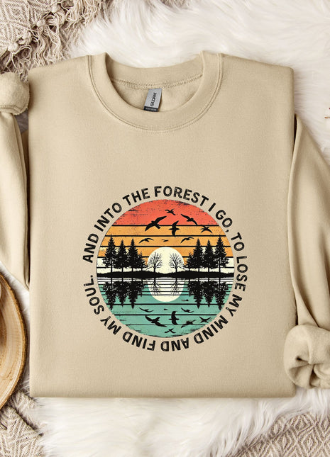 a beige sweatshirt with a graphic design depicting a sunset over a forest, along with the text "And into the forest go I lose my soul" printed on it. The sweatshirt is laid out on a bed with a white blanket, and there are some flowers and a mug nearby.