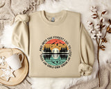 a beige sweatshirt with a graphic design depicting a sunset over a forest, along with the text "And into the forest go I lose my soul" printed on it. The sweatshirt is laid out on a bed with a white blanket, and there are some flowers and a mug nearby.