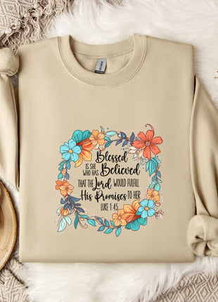 A beige sweatshirt with a floral design and text printed on it, along with a mug, a branch of cotton flowers, and a wooden tray with snowflakes on it, all arranged on a white fur rug.