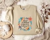 A beige sweatshirt with a floral design and text printed on it, along with a mug, a branch of cotton flowers, and a wooden tray with snowflakes on it, all arranged on a white fur rug.