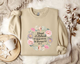 A beige sweatshirt with a floral design and text printed on it, along with a mug, a branch of cotton flowers, and a wooden tray.