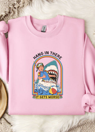 A pink sweatshirt with a graphic design depicting a person in a blue outfit and a shark, along with the text "HANG IN THERE IT GETS WORSE". The sweatshirt is laid out on a bed with a white blanket and a white mug.