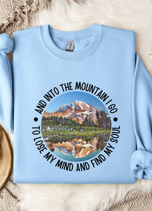 A light blue sweatshirt with a mountain landscape design and the text "And into the mountain I go to lose my mind and find my soul" printed on it, along with a mug and some flowers on a bed.