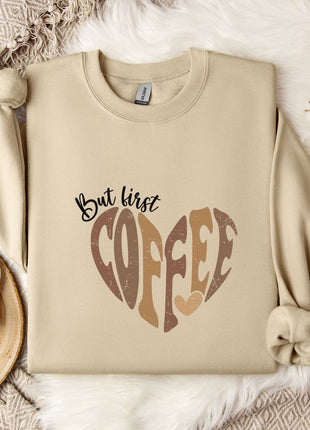 A beige sweatshirt with a heart-shaped design and the text "But first coffee" printed on it, along with a mug and some flowers on a bed.