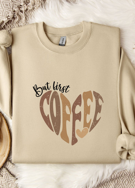 A beige sweatshirt with a heart-shaped design and the text "But first coffee" printed on it, along with a mug and some flowers on a bed.