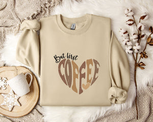 A beige sweatshirt with a heart-shaped design and the text "But first coffee" printed on it, along with a mug and some flowers on a bed.