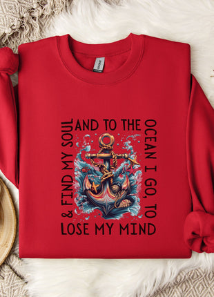 A red sweatshirt with a graphic design of an anchor and the text "AND TO THE OCEAN I GO TO LOSE MY MIND" printed on it, along with a mug and a branch of cotton flowers.