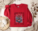 A red sweatshirt with a graphic design of an anchor and the text "AND TO THE OCEAN I GO TO LOSE MY MIND" printed on it, along with a mug and a branch of cotton flowers.