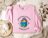 A pink sweatshirt with a graphic design depicting a fire inside a dumpster, accompanied by the text "Emotional Support Dumpster Fire".