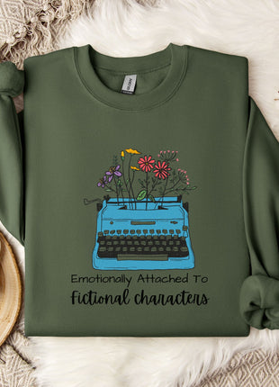 A green sweatshirt with a graphic design of a typewriter filled with colorful flowers, accompanied by the text "Emotionally Attached To Fictional Characters".