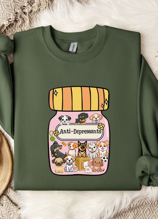 A green sweatshirt with a graphic design of a jar containing various cartoon dogs, along with the text "Ani-Depressants" printed on it. The sweatshirt is placed on a bed with a white blanket and a small white mug.