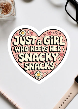 a heart-shaped sticker with the text "Just a girl who needs her snacky snacks" written on it, surrounded by various office supplies such as a watch, a pen, and a pair of glasses.