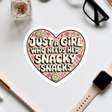 a heart-shaped sticker with the text "Just a girl who needs her snacky snacks" written on it, surrounded by various office supplies such as a watch, a pen, and a pair of glasses.