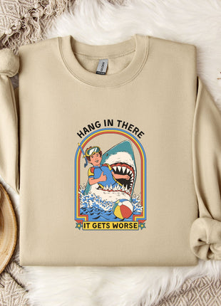 a beige sweatshirt with a graphic design depicting a person holding a shark and the text "HANG IN THERE IT GETS WORSE". The sweatshirt is placed on a bed with a white blanket and a mug.