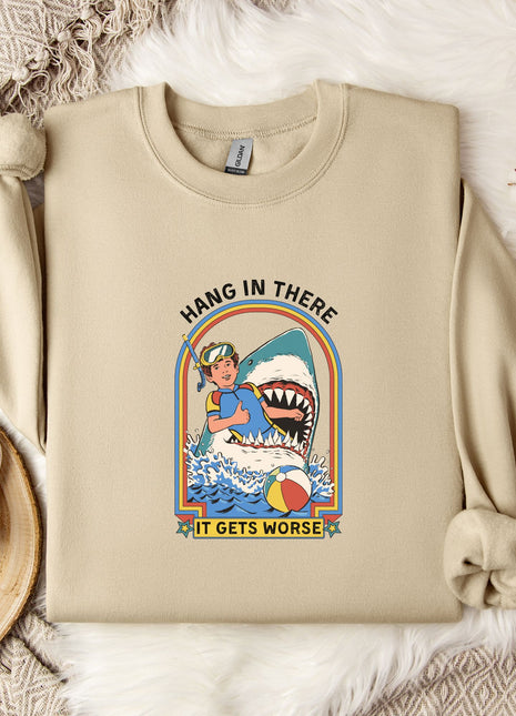 a beige sweatshirt with a graphic design depicting a person holding a shark and the text "HANG IN THERE IT GETS WORSE". The sweatshirt is placed on a bed with a white blanket and a mug.