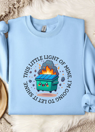 a light blue sweatshirt with a graphic design on it, including a cartoon character and text. The sweatshirt is laid out on a bed with a white blanket and a small white mug.