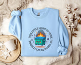 a light blue sweatshirt with a graphic design on it, including a cartoon character and text. The sweatshirt is laid out on a bed with a white blanket and a small white mug.