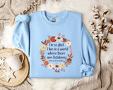 A light blue sweatshirt with a floral wreath design and a quote printed on it, placed on a bed with a white blanket and a white mug.