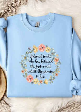 A light blue sweatshirt with a floral design and text printed on it, placed on a bed with a white blanket and a white mug.