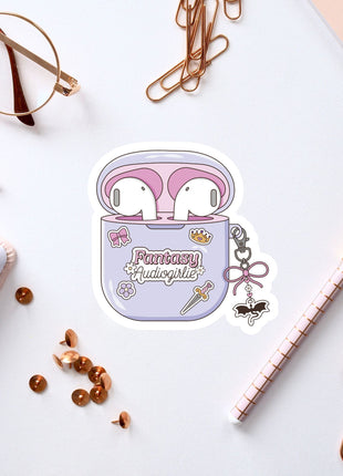 a sticker of a pair of earbuds with the text "Fantasy Audiogirl" on it, surrounded by various office supplies such as pens, a notebook, and a pair of glasses.