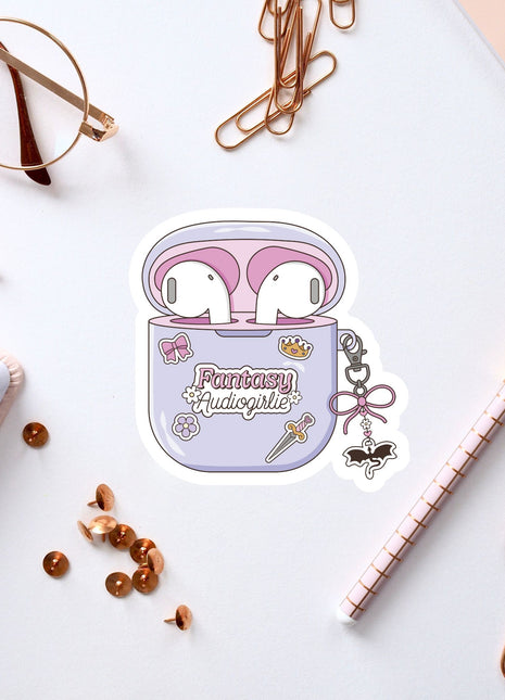 a sticker of a pair of earbuds with the text "Fantasy Audiogirl" on it, surrounded by various office supplies such as pens, a notebook, and a pair of glasses.