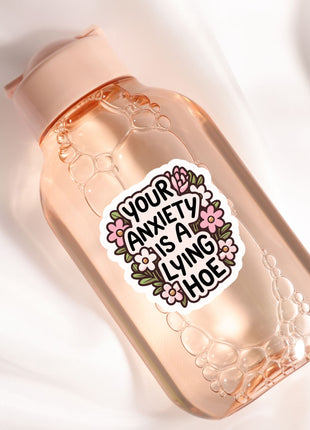 A clear glass bottle with a pink cap, containing a liquid and surrounded by bubbles. The bottle has a sticker on it with the text "Your anxiety is a living hoe" in a floral design.