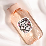 A clear glass bottle with a pink cap, containing a liquid and surrounded by bubbles. The bottle has a sticker on it with the text "Your anxiety is a living hoe" in a floral design.