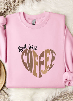 A pink sweatshirt with a heart-shaped design and the text "But first coffee" printed on it, along with a mug and some flowers on a bed.