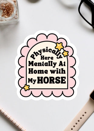 a sticker with a humorous text that reads "Physically Here Mentally At Home With My Horse" surrounded by various office supplies and accessories such as a pair of glasses, a pen, and a watch.