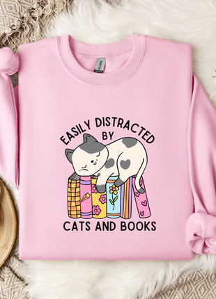 A pink sweatshirt with a graphic design of a cat sitting on a stack of books, accompanied by the text "EASILY DISTracted BY CATS AND BOOKS".