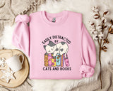 A pink sweatshirt with a graphic design of a cat sitting on a stack of books, accompanied by the text "EASILY DISTracted BY CATS AND BOOKS".