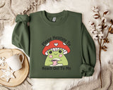 a green sweatshirt with a mushroom character and text, along with a mug, a white mug, and some white flowers on a white fur rug.