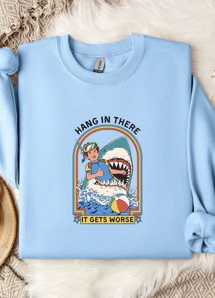 A light blue sweatshirt with a graphic design depicting a shark and the text "HANG IN THERE IT GETS WORSE" is displayed on a bed with a white blanket and a mug.