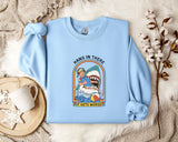 A light blue sweatshirt with a graphic design depicting a shark and the text "HANG IN THERE IT GETS WORSE" is displayed on a bed with a white blanket and a mug.