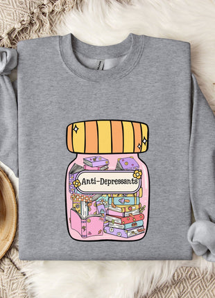 A gray sweatshirt with a colorful graphic of a jar containing various objects, including books and a teapot, along with the text "Ani-Depressants" printed on it. The sweatshirt is laid out on a bed with a blanket and a mug, and there are some white flowers nearby.
