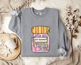 A gray sweatshirt with a colorful graphic of a jar containing various objects, including books and a teapot, along with the text "Ani-Depressants" printed on it. The sweatshirt is laid out on a bed with a blanket and a mug, and there are some white flowers nearby.