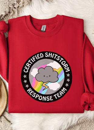 a red sweater with a graphic design on it, placed on a white fur rug. The design includes a cloud with a lightning bolt and the text "Certified Shitstorm Response Team".