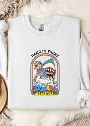 A white sweatshirt with a graphic design of a person surfing with a shark, accompanied by the text "HANG IN THERE IT GETS WORSE". The sweatshirt is laid out on a bed with a white blanket and a white mug.