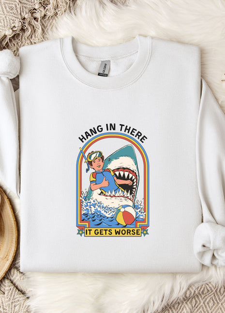 A white sweatshirt with a graphic design of a person surfing with a shark, accompanied by the text "HANG IN THERE IT GETS WORSE". The sweatshirt is laid out on a bed with a white blanket and a white mug.