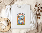 A white sweatshirt with a graphic design of a person surfing with a shark, accompanied by the text "HANG IN THERE IT GETS WORSE". The sweatshirt is laid out on a bed with a white blanket and a white mug.