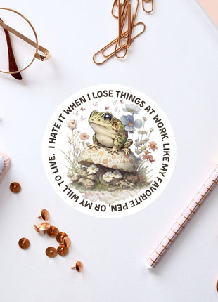 a sticker with a frog sitting on a mushroom, surrounded by various office supplies such as pens, paper clips, and a pair of glasses.