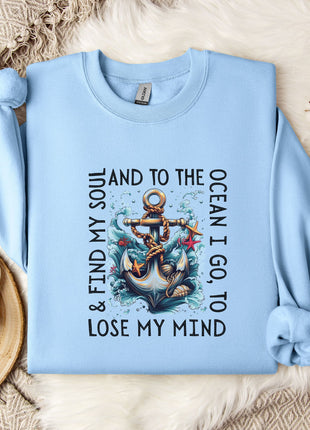 A light blue sweatshirt with a graphic design of an anchor and waves, along with the text "& FIND MY SOUL AND TO THE OCEAN I GO TO LOSE MY MIND" printed on it. The sweatshirt is laid out on a bed with a white blanket and a mug, and there are some white flowers nearby.